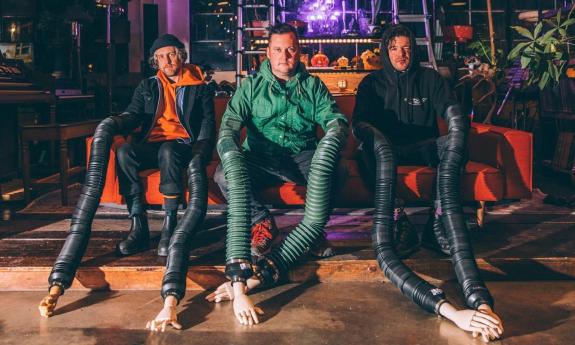 Bandmates from Modest Mouse sit on an orange couch wearing strange black and green clothing with elongated arms.