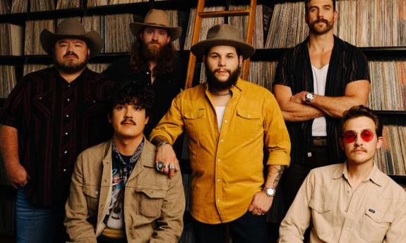 Bandmates from Flatland Cavalry huddle together and pose in front of a large book shelf with a ladder. 