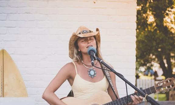 Musician Lucy Spotts in cowboy hat, with guitar, in front of white brick wall