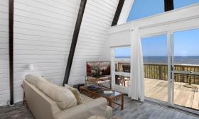 A large floor to ceiling window and glass door face the ocean in a beach house offered by Florida Rentals. Comfortable furniture and a TV in the foreground