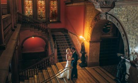 A couple embraces at the marble mosaic entrance to the Ponce Hall Dining Room, backed by Tiffany stained glass windows and warm lighting..