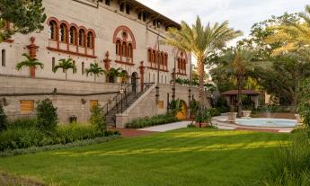 A photo of a landscaped garden and lawn, with water feature and palm trees with stairs to an historic building with red-trimmed windows