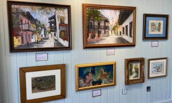 A white slatboard wall holding six fine art paintings that portray city streets, musicians, and Florida landscapes