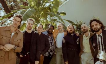 Bandmates from The Revivalists huddle together and pose in front of an oversized plant.