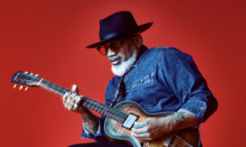 Toronzo Cannon wears black glasses while strumming his guitar in front of a red backdrop. 