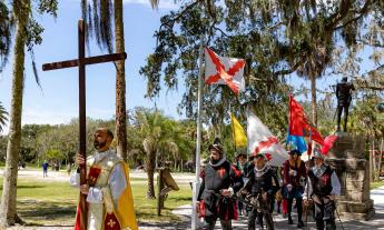 A priest holding a wooden cross leads men dressed as Spanish soldiers from the 1500s