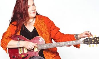 Carolyn Wonderland smiles while holding a guitar in front of a white backdrop.