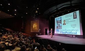 Two people stand on a stage in a darkened autorium, in front of a large movie screen