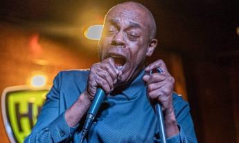 Michael Winslow wears a blue shirt while speaking with a microphone pressed to to his lips.