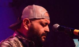 Jon Q., in profile, wearing a basball cap and facing the microphone