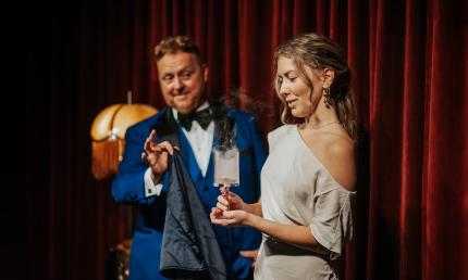 A magician in front of a velvet curtain, performs magic with the aide of a woman from the audience