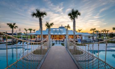 A bridge over a pool, leading to a clubhouse framed by palm trees at sunset